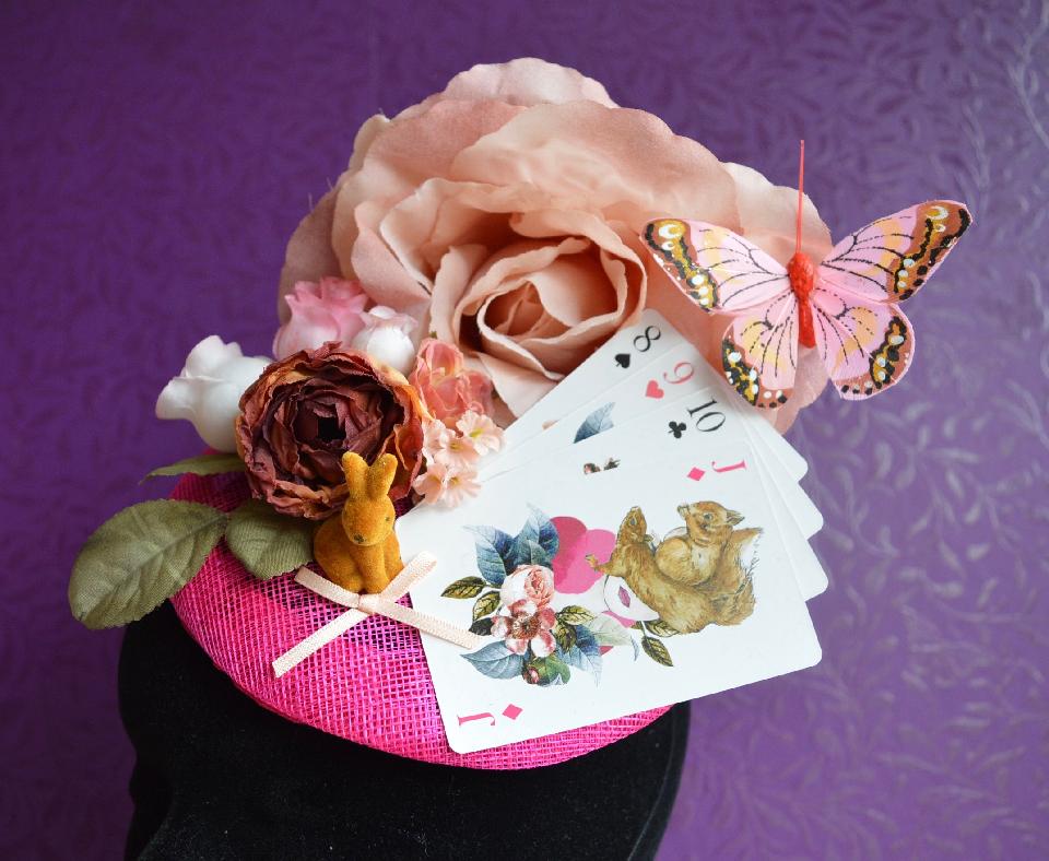 IMAGE - Pink sinamay fascinator with pink flowers, playing cards, bunny and butterfly. Finished with a pink bow. Fixes to hair with a comb.