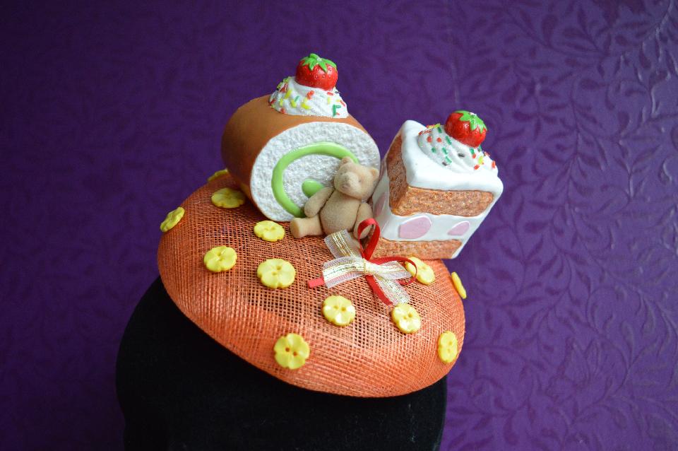 IMAGE - Orange sinamay fascinator with two pieces of cake and a bear. Decorated with bows and yellow flower buttons. Fixes to hair with a comb.