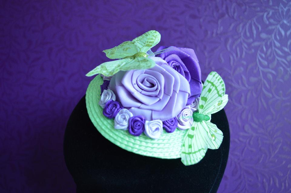 IMAGE - Green straw fascinator decorated with purple and lilac ribbon roses and green butterflies. Fixes to hair with a comb.